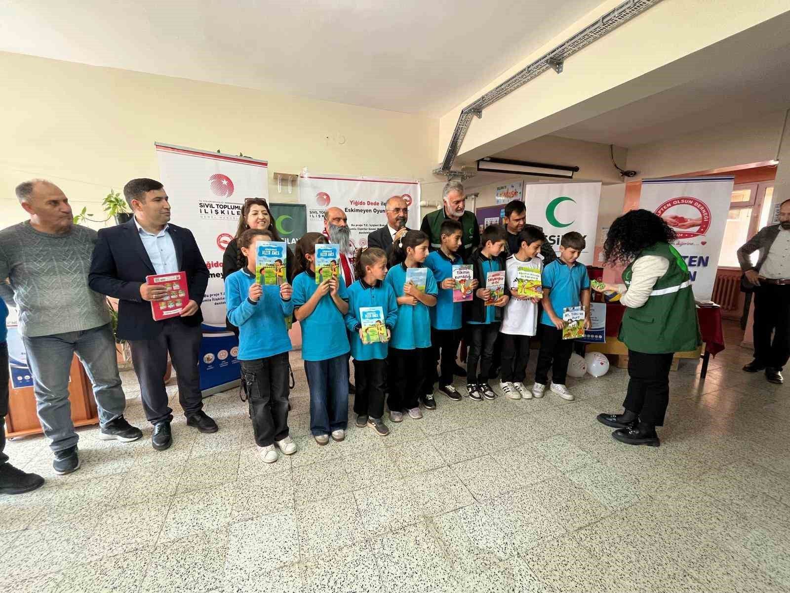 Bir grup çocuk, ellerindeki kitaplarla yetişkinlerin yanında kapalı salonda yan yana duruyor.