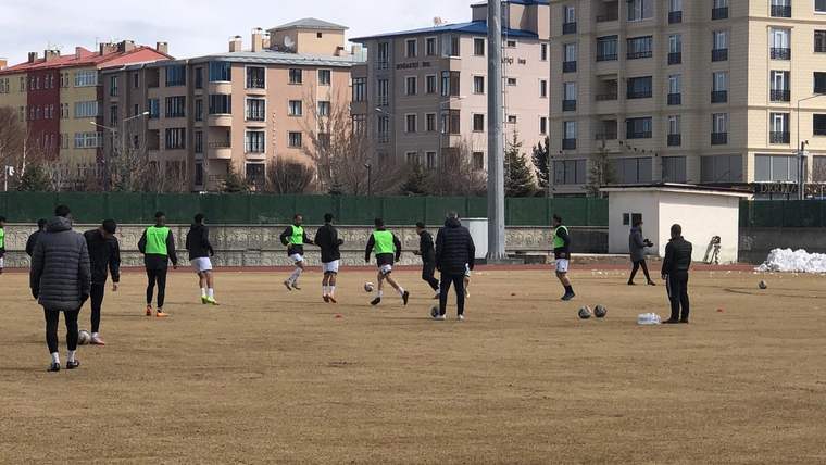 Futbol sahasında yeşil formalı oyuncular top sürerken, kenarda koyu montlu kişiler onları izliyor.