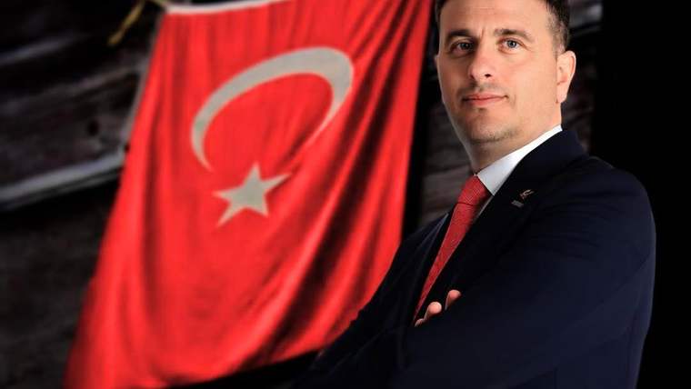 Kırmızı bir Türk bayrağının önünde, takım elbise giymiş bir kişi duruyor.