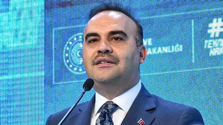 Takım elbiseli bir kişi, yakasında Türk bayrağı rozetiyle mikrofon karşısında konuşurken görülüyor.