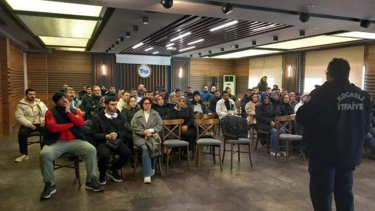 Toplantı salonunda oturan kalabalık bir gruba, sırtında itfaiye yazısı bulunan kişi konuşuyor.