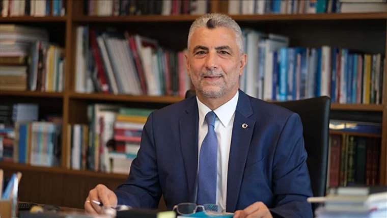 Kitaplık önünde takım elbiseli bir kişi, masada otururken ellerini ve gözlüğünü önünde tutuyor.