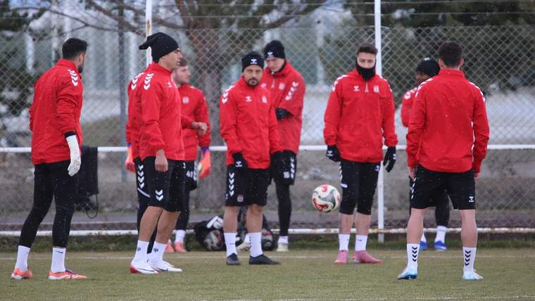 Kırmızı antrenman giysileri giymiş futbolcular, antrenman sahasında toplanmış ve topa odaklanmış durumda.