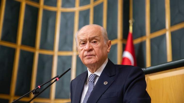 Kürsüde konuşan bir kişi, arkasında Türk bayrağı ve ahşap bir panel var.