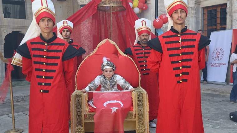 Sünnet kıyafeti giymiş bir çocuk, tahtta oturuyor; yanında iki kişi geleneksel kıyafetler içinde duruyor.