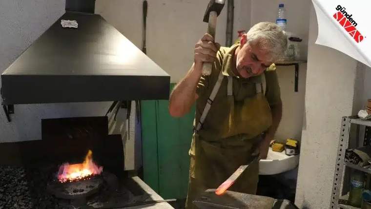 Önlük giyen bir kişi, örs üzerindeki kızgın metal parçaya çekiçle vururken, ocak alevi yanında görülüyor.