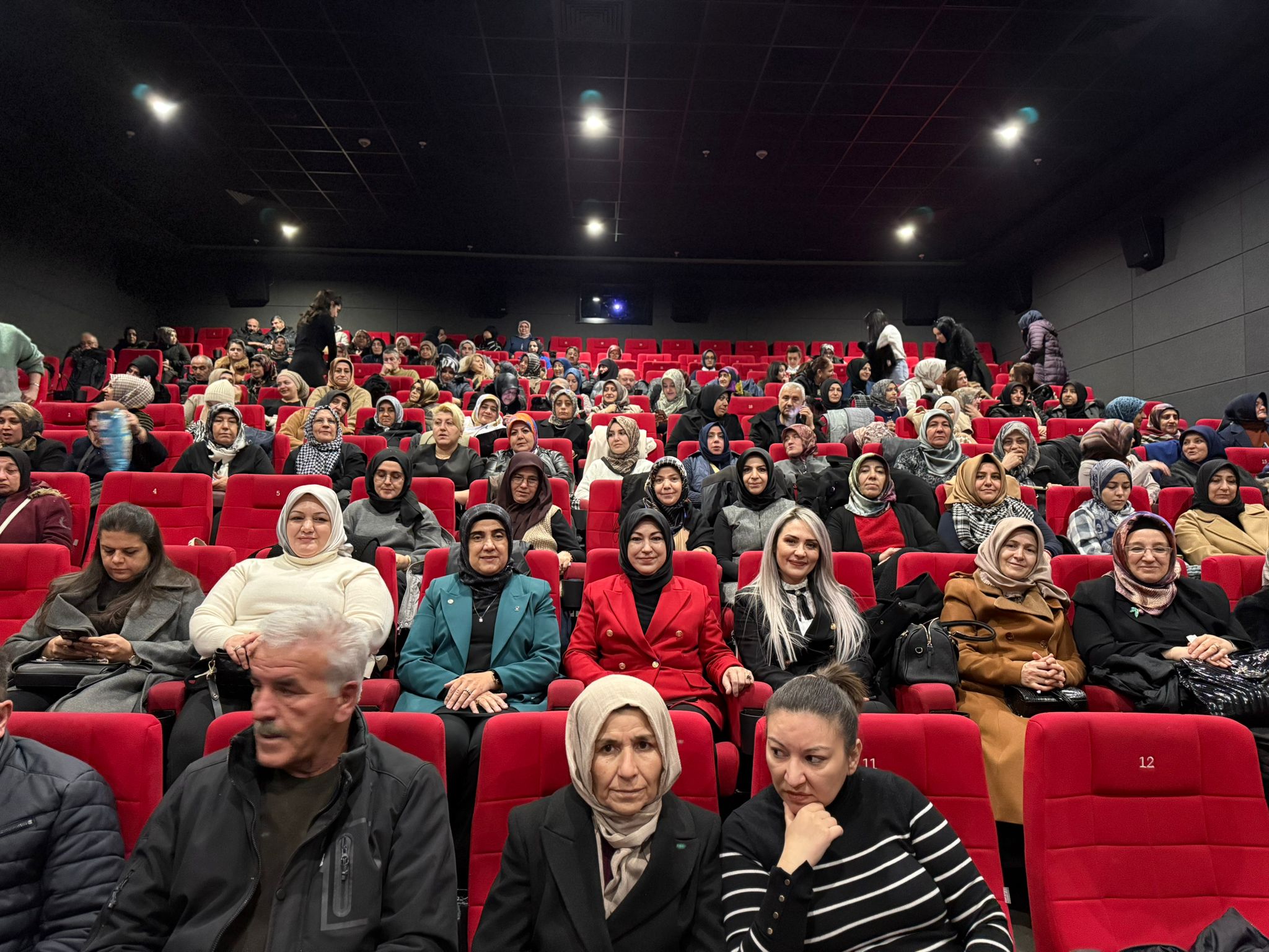 Kırmızı koltuklarda oturan kalabalık bir izleyici grubu, sinema salonunda film izliyor.