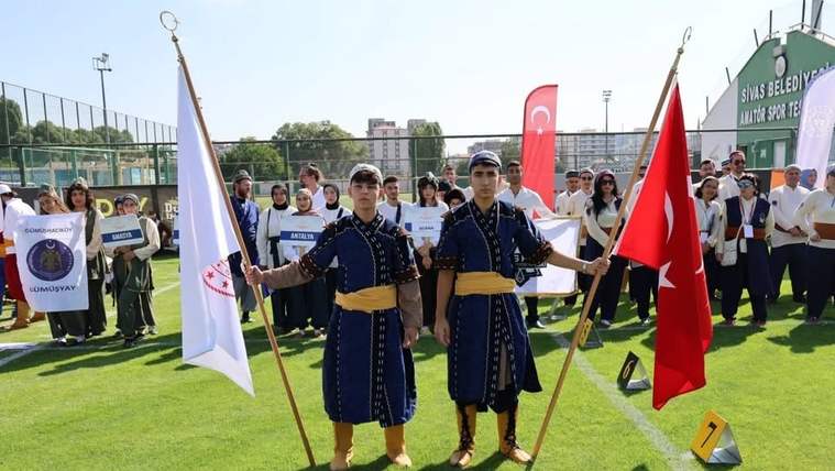 Saha üzerinde geleneksel kıyafetli iki genç, beyaz ve Türk bayraklarını taşırken arka planda sporcular sıralanıyor.
