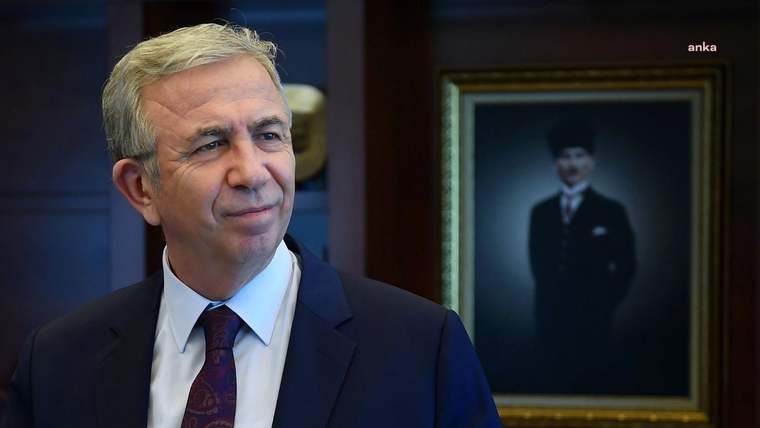 Takım elbiseli bir kişi, arkasındaki çerçeveli üniformalı portreyle birlikte görülüyor.