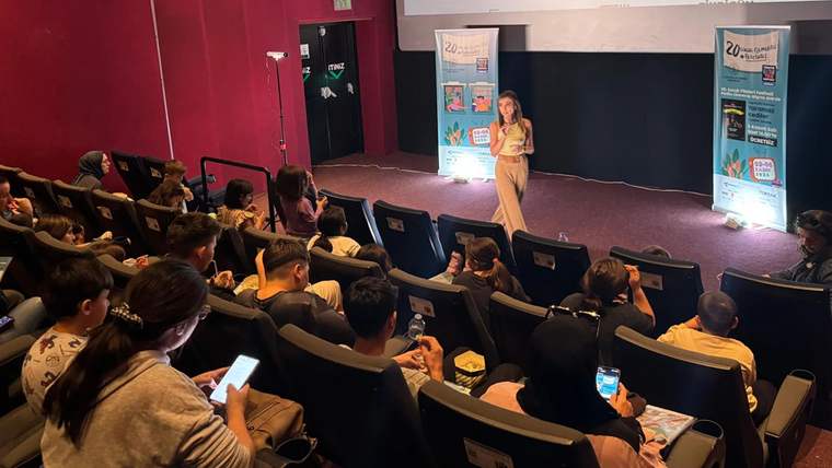 Bir sinema salonunda, çocuklara yönelik bir etkinlikte konuşma yapan bir kadın, dinleyici olarak oturan çocuklar ve aileleri var.
