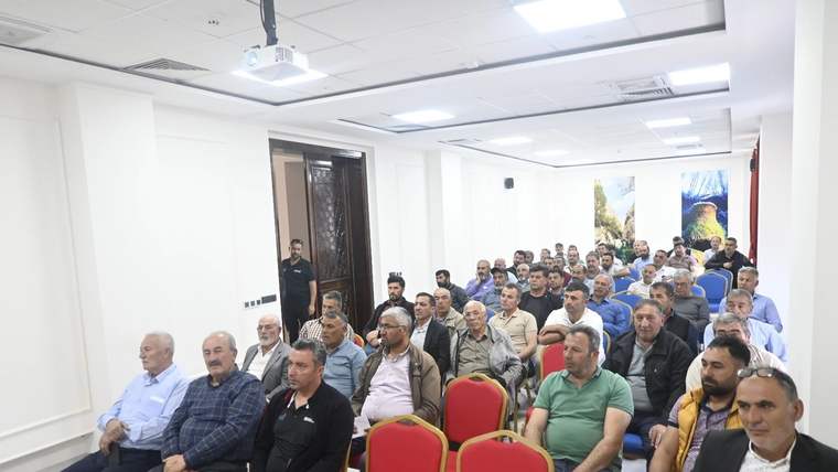Toplantı salonunda sıralar halinde oturan çok sayıda kişi ve ön bölümde boş kırmızı sandalyeler görülüyor.
