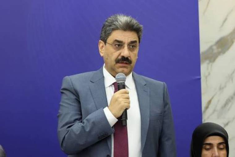 Kürsüde mikrofonla konuşan bıyıklı bir kişi, yanında başörtülü bir kadınla birlikte görülüyor.