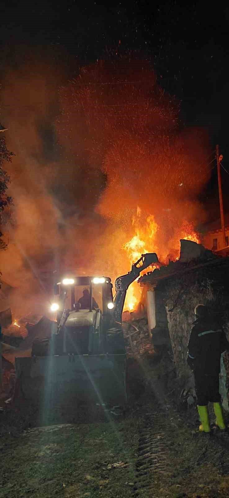Gece karanlığında alevler içindeki yapının önünde iş makinesi ve hortum taşıyan bir kişi görülüyor.