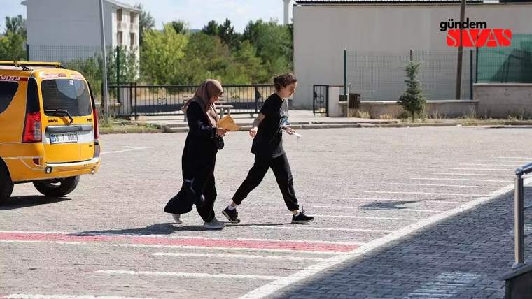 İki kişi, bir otoparkta yürüyerek ilerliyor. Bir kişi elinde belgeler tutuyor.