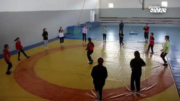 Spor salonunda genç kızlar, renkli minderlerle çevrili alanda top oyununa katılıyor.