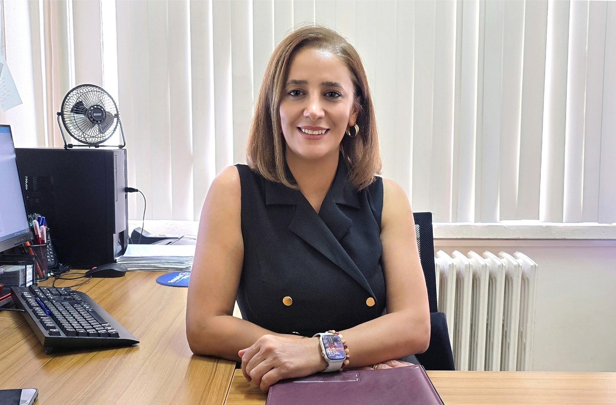 Bir masa başında oturan, gülümseyen bir kadın, önünde bilgisayar ve dosya var.