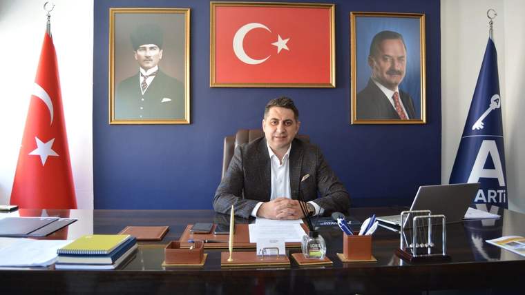 Makam masasının arkasında oturan bir kişi, Türk bayrağı ve iki portreyle çevrili çalışma odasında görülüyor.