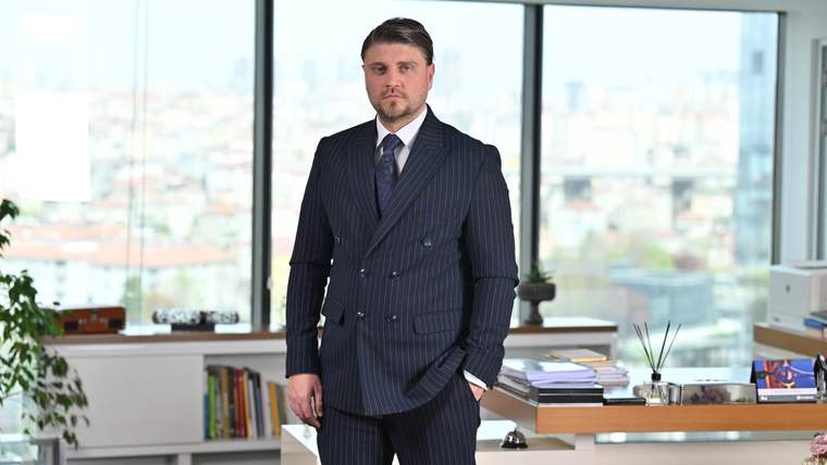 Ofis ortamında koyu renk takım elbiseli bir kişi, çalışma masasının yanında ayakta duruyor.
