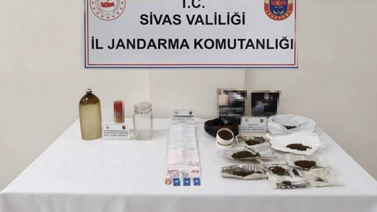 Masada çeşitli paketler, kavanoz ve şişeler; arkada Sivas Valiliği Jandarma Komutanlığı yazılı pano bulunuyor.