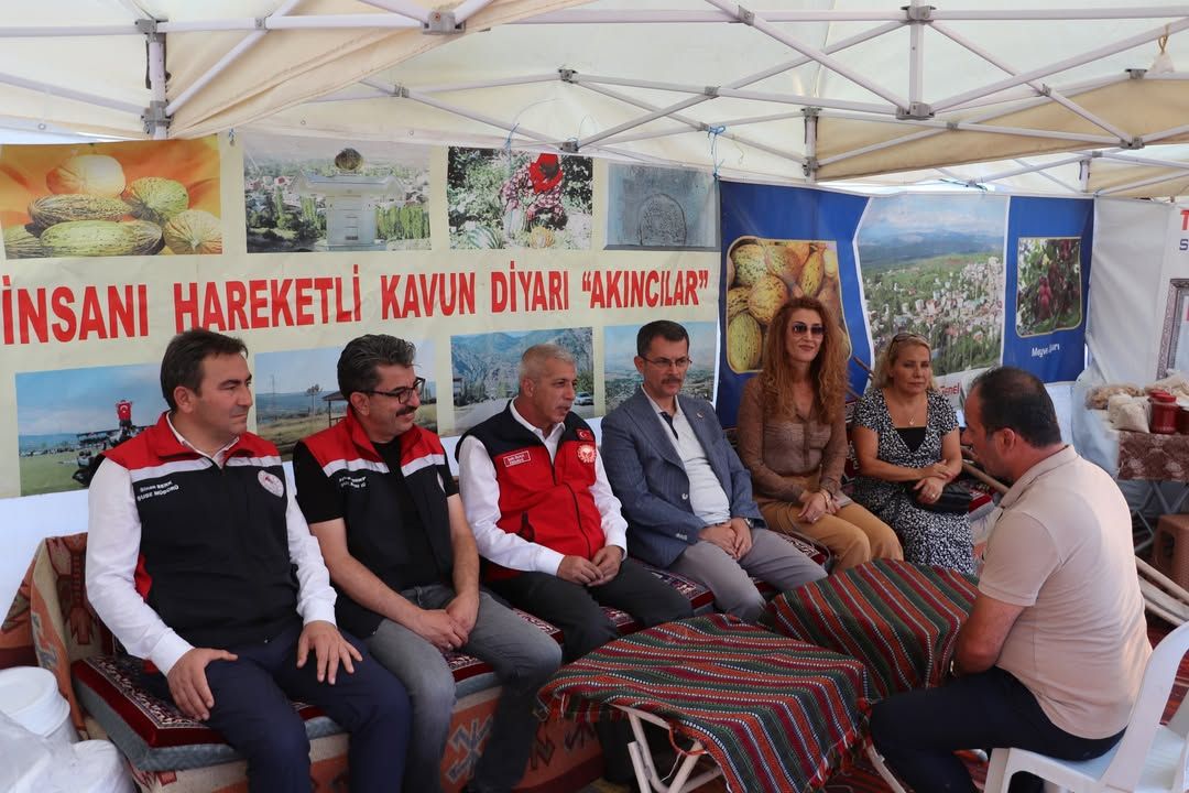 Çadır altında oturan altı kişi, karşılarındaki bir kişiyle sohbet ediyor; arkada ürün ve manzara afişleri bulunuyor.