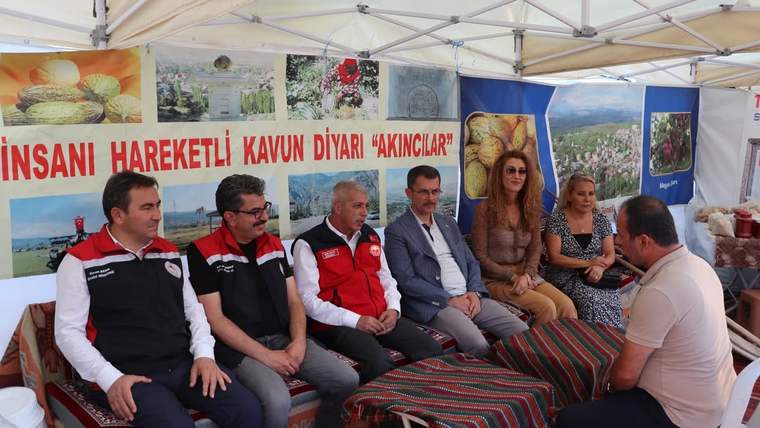 Çadır altında oturan altı kişi, karşılarındaki bir kişiyle sohbet ediyor; arkada ürün ve manzara afişleri bulunuyor.