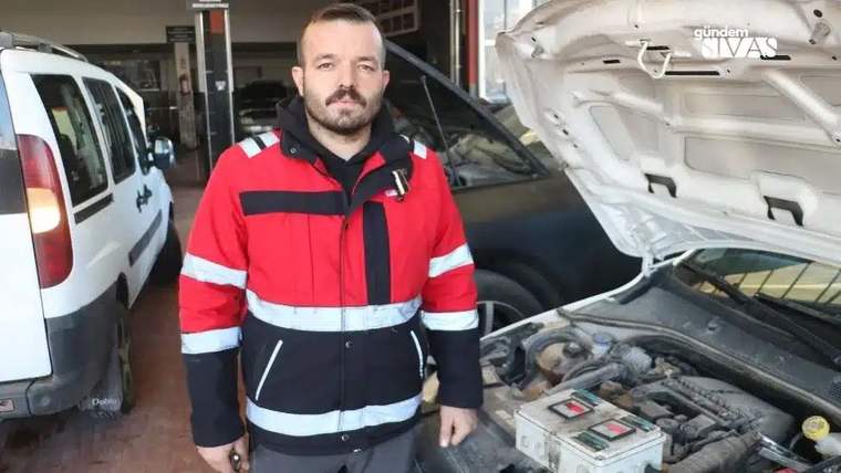 Kırmızı iş kıyafetli bir kişi, kaputu açık otomobilin motoru ve aküsü yanında duruyor.