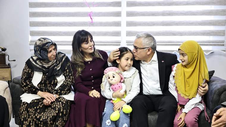 Bir kanepede oturan beş yetişkin ve bir çocuk; ortadaki çocuk kucağında oyuncak bebek tutuyor.