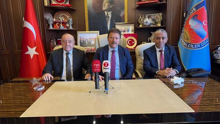 Üç kişi, masanın etrafında oturmuş, arka planda Türk bayrağı ve fotoğraflar var.