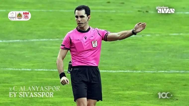 Pembe formalı bir futbol hakemi, yeşil saha üzerinde sağ kolunu yana doğru uzatıyor.