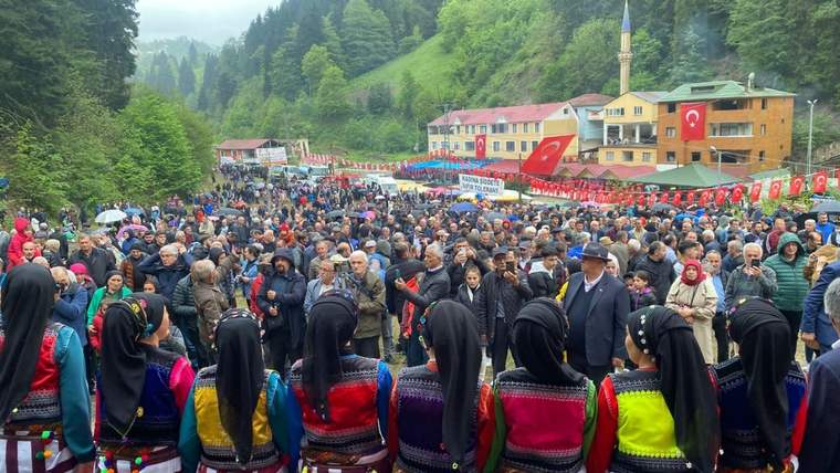 Geleneksel kıyafetli kadınların önünde, ormanlık vadide Türk bayraklarıyla çevrili kalabalık topluluk görülüyor.