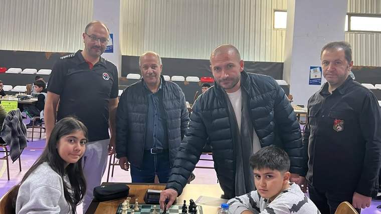 Satranç salonunda iki çocuk masadaki tahtada oynarken dört yetişkin arkalarında duruyor.