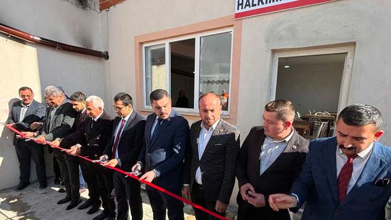 Bir binanın önünde takım elbiseli kişiler kırmızı kurdele keserken, sağda makasların bulunduğu tepsi tutuluyor.
