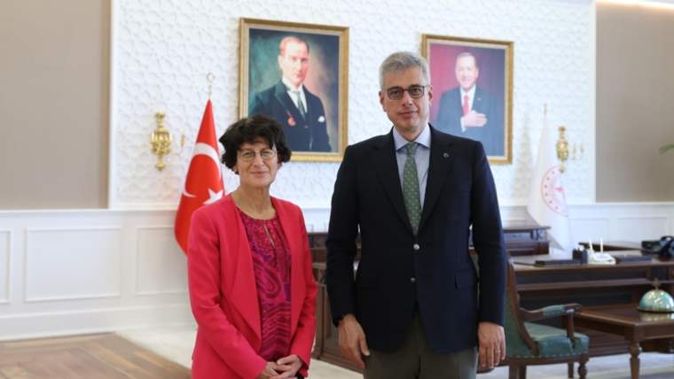 Bir odada kırmızı ceketli bir kadın ve takım elbiseli bir erkek, Türk bayrağı ve duvar portreleri önünde duruyor.