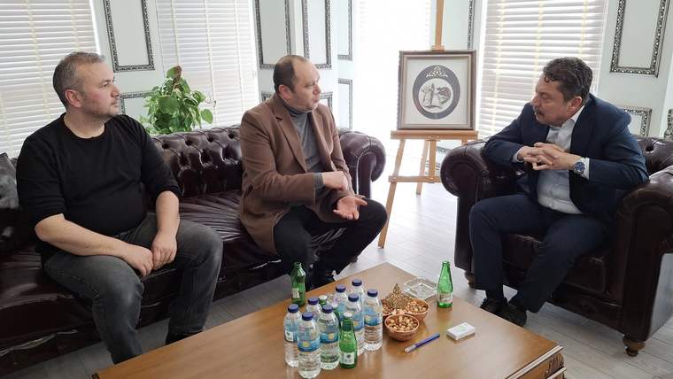 Bir odada koltuklarda oturan üç erkek, üzeri içecek ve atıştırmalıklarla dolu sehpanın çevresinde konuşuyor.