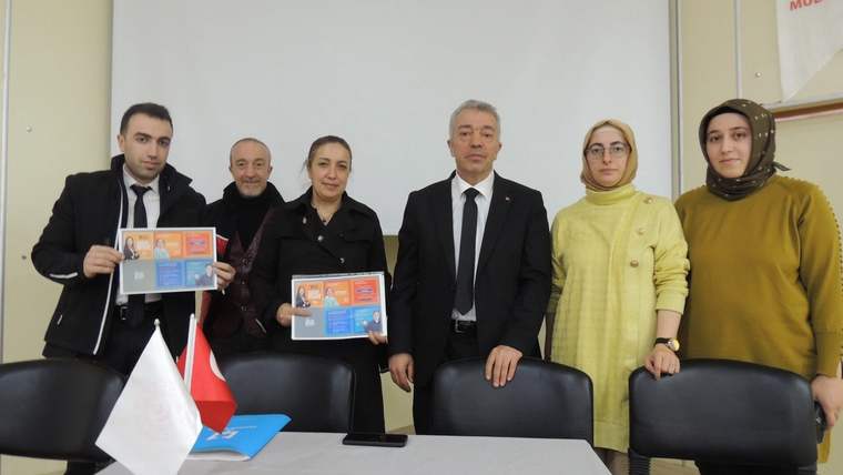 Toplantı odasında altı kişi duruyor; iki kişi ellerinde bilgilendirme broşürü tutuyor, masada Türk bayrağı bulunuyor.