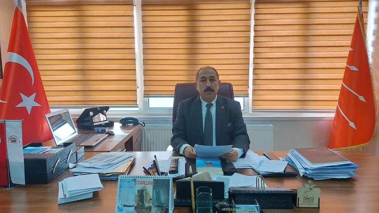 Masanın arkasında takım elbiseli bir kişi, önünde evraklar ve bilgisayar bulunan odada oturuyor.