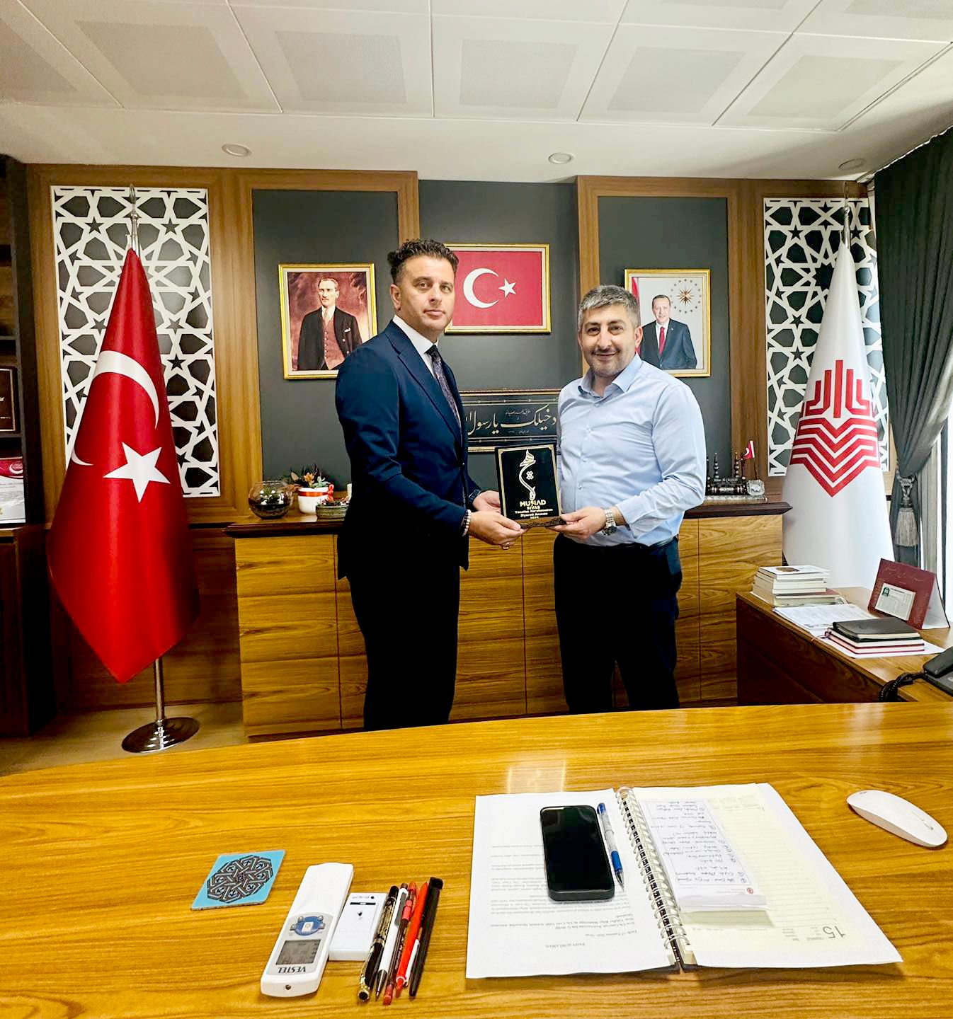 MÜSİAD temsilcisi ve Vakıflar Bölge Müdürü, bir plaket tutarak masanın önünde duruyor.