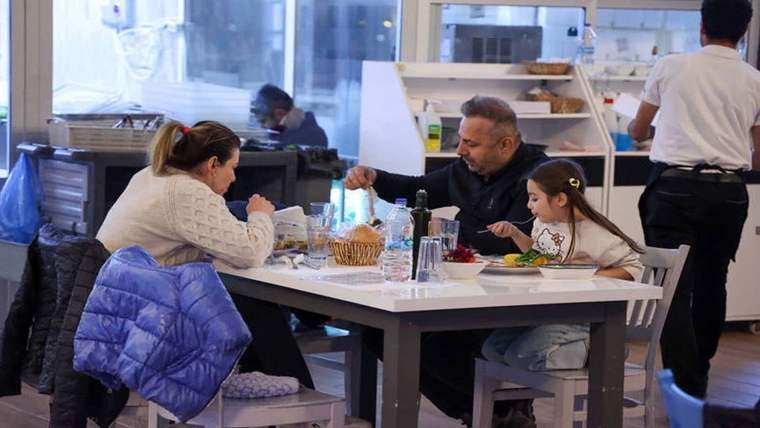 Lokantada bir adam, kadın ve küçük kız çocuğu masada yemek yerken; arkada çalışanlar görülüyor.