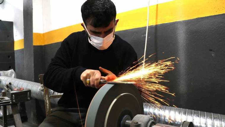 Maskeli bir kişi, atölyede döner taşlama makinesine metal bir bıçak tutarken kıvılcımlar çıkıyor.
