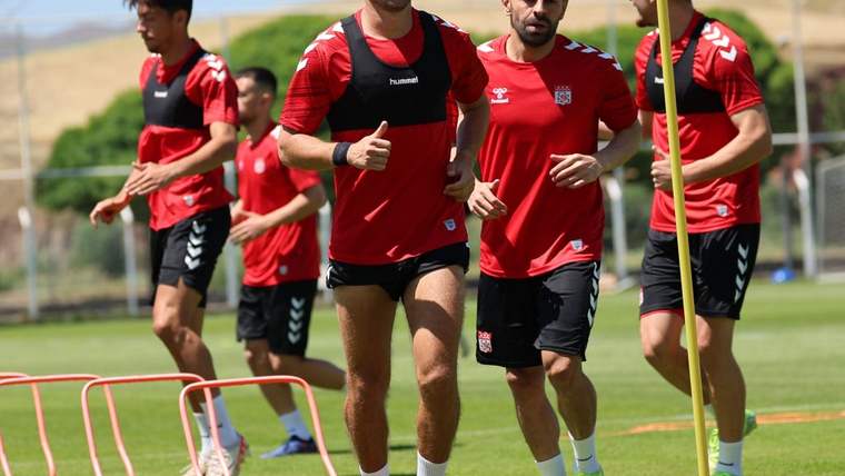 Kırmızı formalar giyen futbolcular, sahada antrenman sırasında engeller ve slalom direkleri arasında koşuyor.