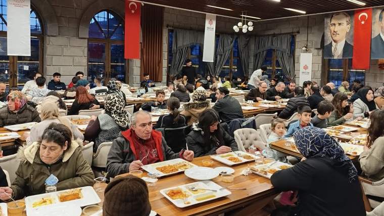 Geniş bir salonda çok sayıda kişi uzun masalarda yemek yerken duvarlarda Türk bayrağı ve portre görülüyor.