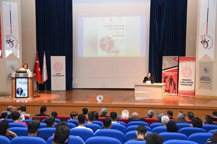 Konferans salonunda konuşmacı kürsüde yer alırken izleyiciler mavi koltuklarda oturuyor, ekranda sunum görünüyor.