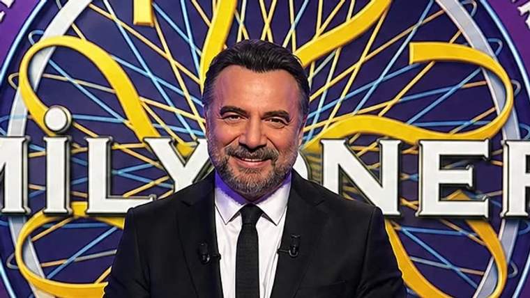 Gülümseyen bir sunucu, arka planda 'Milyoner' yazılı bir afişin önünde duruyor.