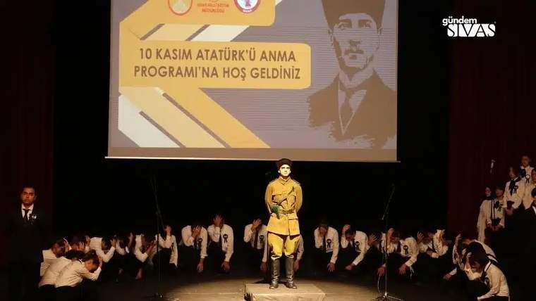 Sahnede sarı kostümlü bir çocuk, çevresinde öğrenciler ve arkada 10 Kasım Atatürk’ü Anma yazılı perde var.
