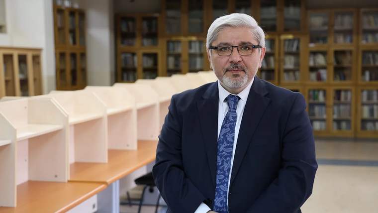 Kütüphane ortamında, takım elbise giymiş bir kişi, masanın önünde duruyor.