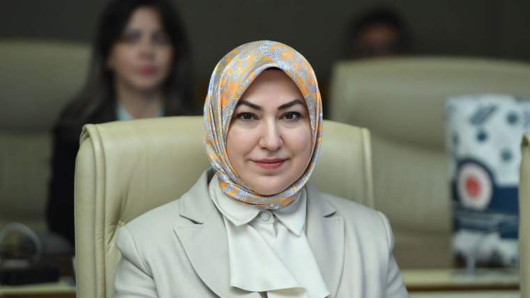 Bir kadın, krem rengi bir ceket giymiş, arka planda başka kişiler var. Kadının başında desenli bir başörtüsü var.