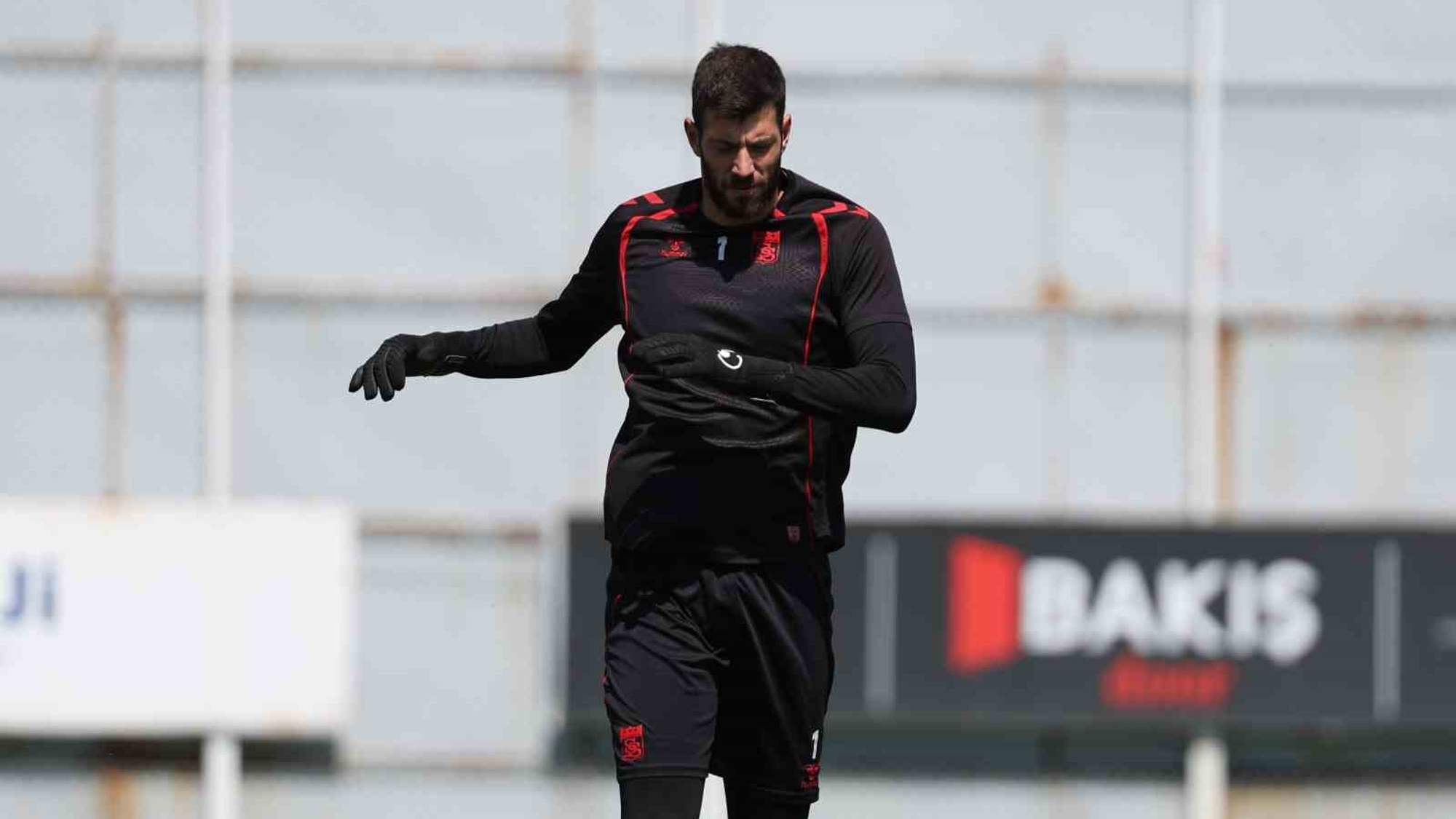 Antrenman yapan bir futbolcu, siyah antrenman kıyafeti giymekte ve eldiven takmakta.
