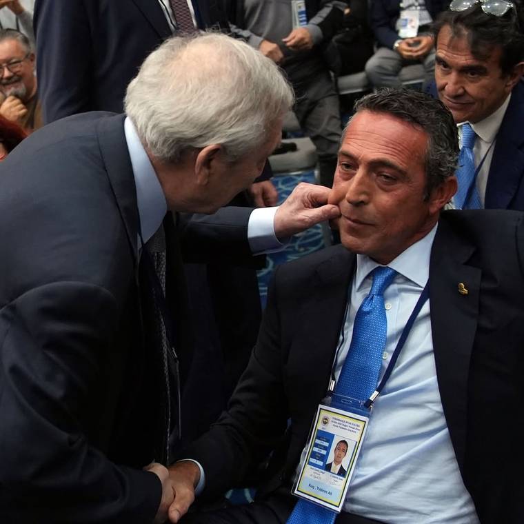 Takım elbiseli iki erkekten biri otururken diğeri elini yanağına uzatıyor; arkada insanlar görülüyor.