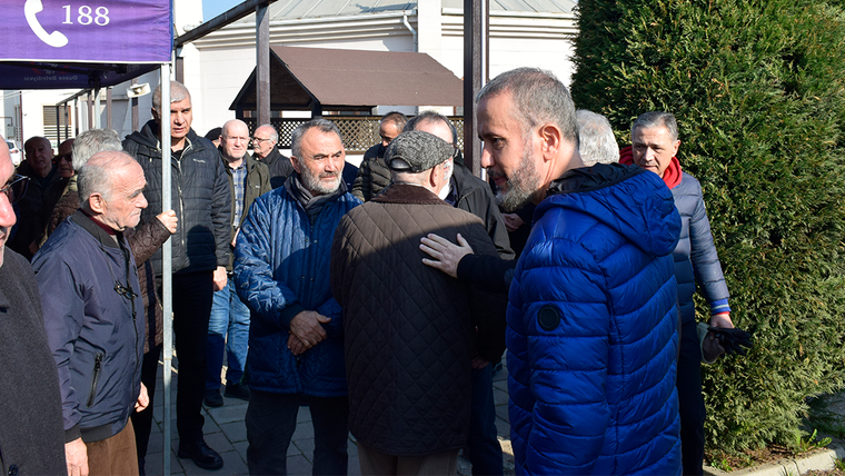 Açık havada bir araya gelen erkekler arasında, mavi montlu bir kişi yaşlı bir erkeğin sırtına elini koyuyor.