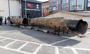 Büyük, paslanmış metal boru, kaldırım kenarında metal ayaklar üzerinde uzanıyor; arkada camlı bina bulunuyor.